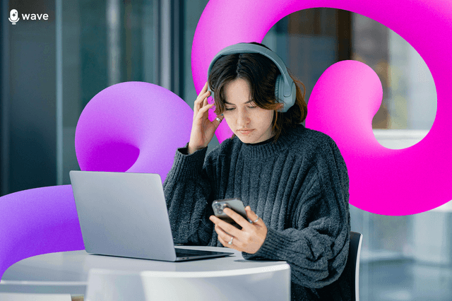 A person sitting at a laptop listening to headphones and looking at their phone